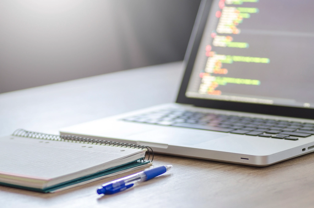Laptop mit Programmcode neben Notizblock und Stift