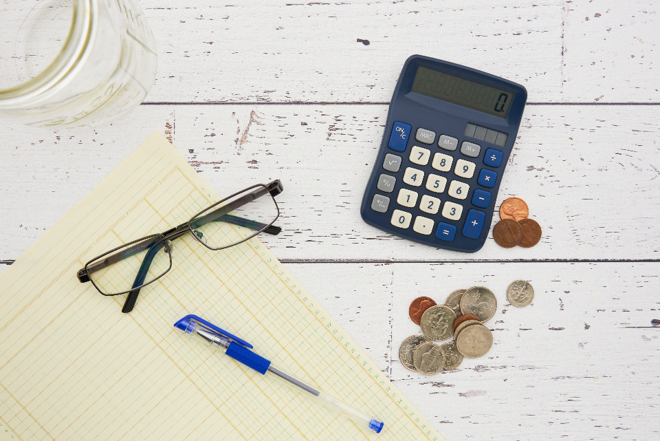 Taschenrechner, Brille, Münzen und Notizblock auf einem Schreibtisch zum Thema Unternehmensfinanzierung